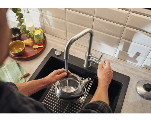 Osoba plní kanvicu v dreze vodovodným kohútikom