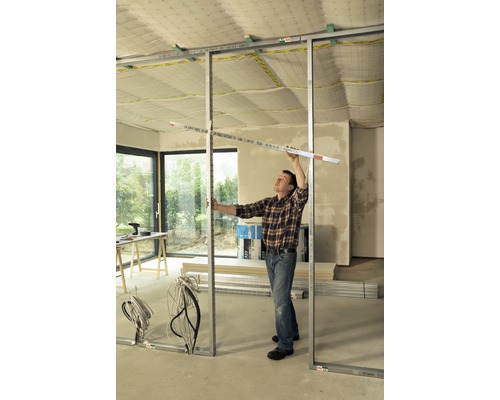 Muž montuje sadrokartónovú stenu s kovovými profilmi a meracím nástrojom v miestnosti.