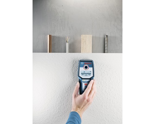 Ruka drží detektor Bosch na stene. Na poličke v pozadí je medené potrubie, elektrický kábel, kus dreva a výstuž.