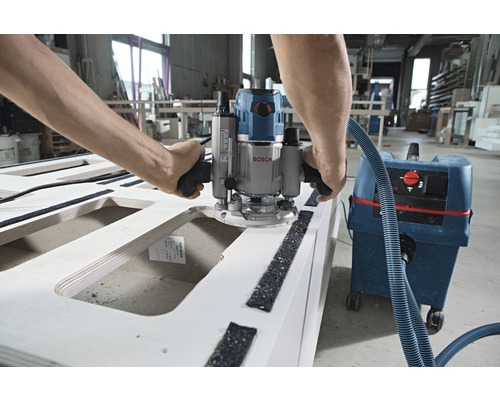 Muž pracuje s hornou frézou na drevenom obrobku, pripojenou k vysávaču.