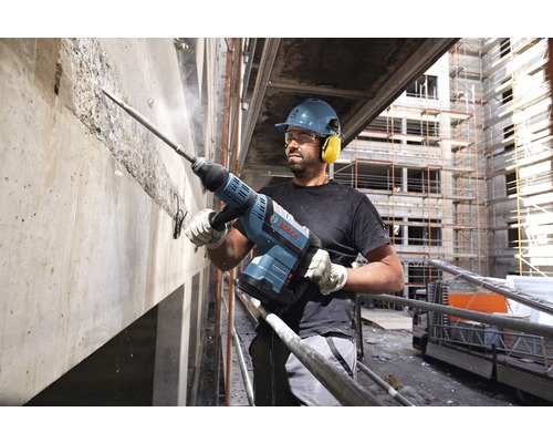 Stavebný robotník s ochrannou helmou a chráničmi sluchu pracuje s demolačným kladivom na betónovom múre. Logo Bosch.