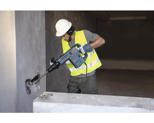 Muž s ochrannou prilbou a bezpečnostnou vestou používa zbíjacie kladivo na stenu.