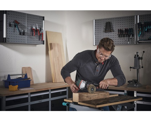 Muž pracuje s drôtenou kefou a uhlovou brúskou na obrobku pred pracovným stolom a stenou s náradím.