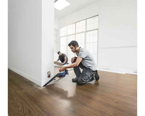 Dvaja remeselníci kladú laminátovú podlahu s ponornou pílou vo svetlej miestnosti.