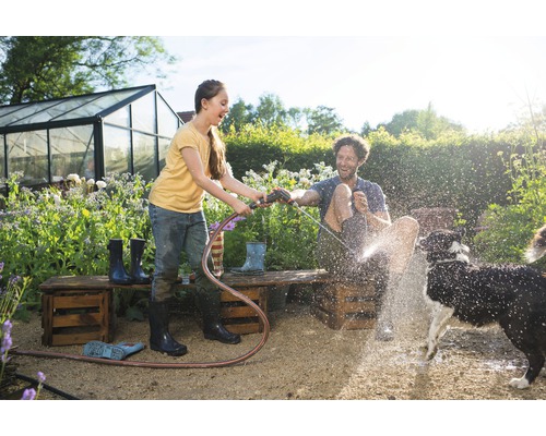Mladá žena drží záhradnú hadicu a strieka muža a psa v záhrade. V pozadí je skleník.