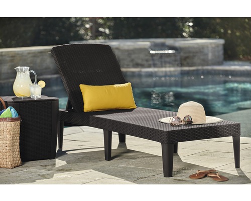 Scéna s tmavým záhradným lehátkom, odkladacím stolíkom, karafou s nápojom, žltým vankúšom, slameným klobúkom a slnečnými okuliarmi pri bazéne.