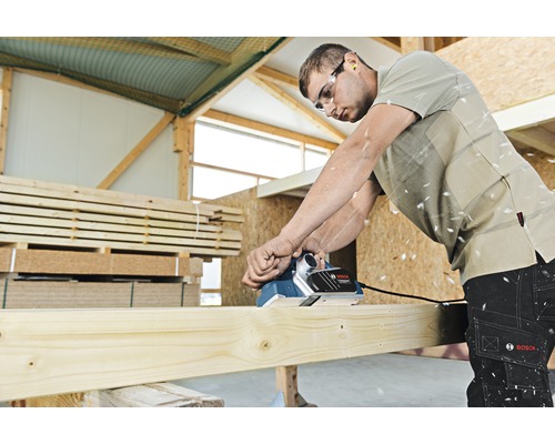 Muž hobľuje drevenú dosku elektrickým hoblíkom v dielni. Muž má na sebe chrániče sluchu a ochranné okuliare. Logo Bosch.