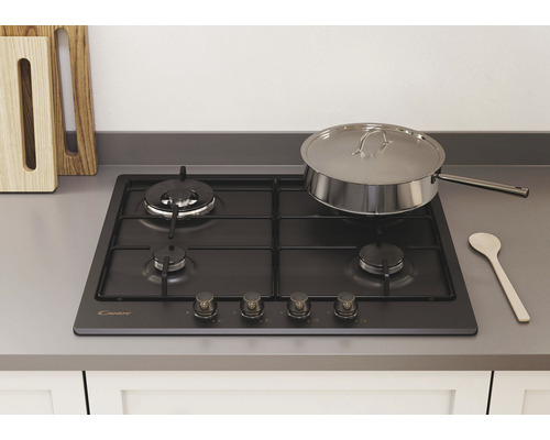 Čierny plynový sporák Candy so štyrmi horákmi, nerezovým hrncom a drevenou lyžicou na pracovnej doske.