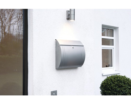 Strieborná poštová schránka na bielej stene vedľa lampy a okna