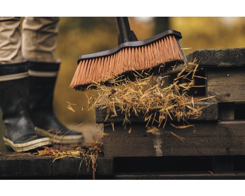 Osoba zametá lístie a záhradný odpad z drevenej terasy pouličnou metlou.