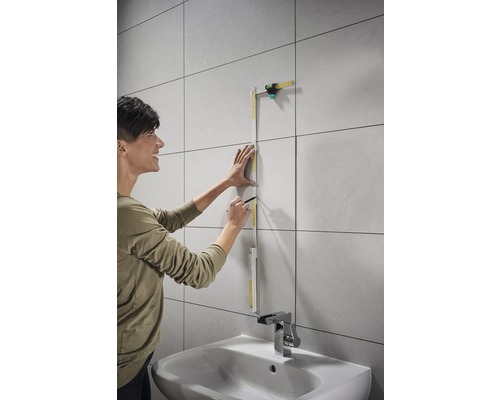 Žena meria obkladanú stenu skladacím metrom a ceruzkou v kúpeľni nad umývadlom.