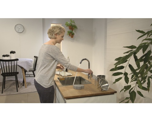 Svetlá kuchyňa so ženou pri umývadle, kohútik, drez a kuchynské náčinie.