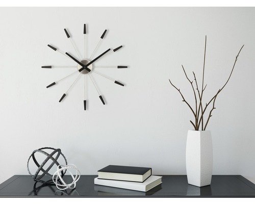 Dekoratívna scéna s nástennými hodinami, vázou, konármi, knihami a ozdobnými guľami na stole.