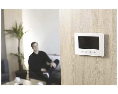 Interiérový záber obývačky s mužom na pohovke a bielym monitorom dverového telefónu na stene.