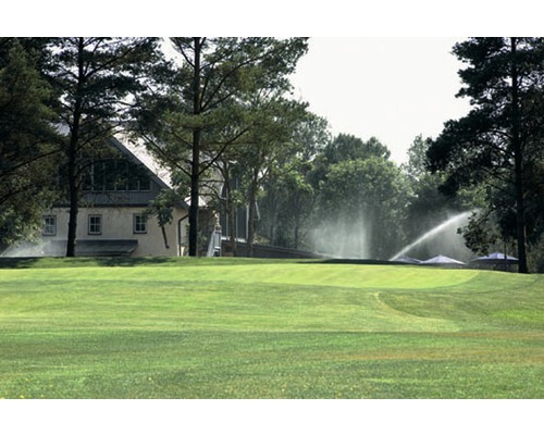 Zavlažovací systém na zelenom trávniku so stromami a budovou v pozadí