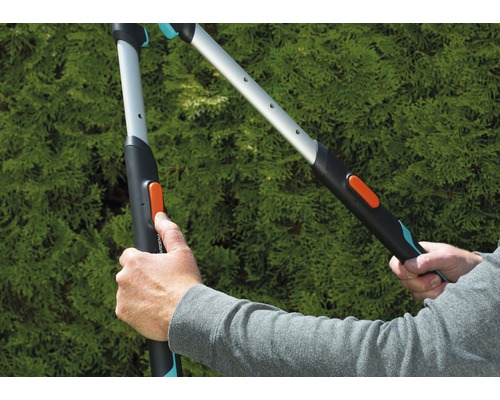 Dve ruky ovládajú záhradné nožnice pred zeleným kríkom.