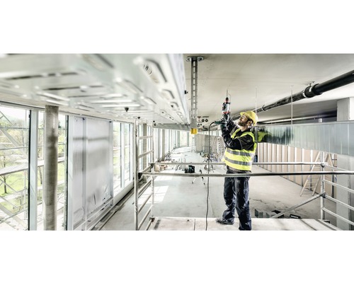 Stavebný robotník na lešení pracuje s vŕtacím kladivom na stropnej inštalácii.