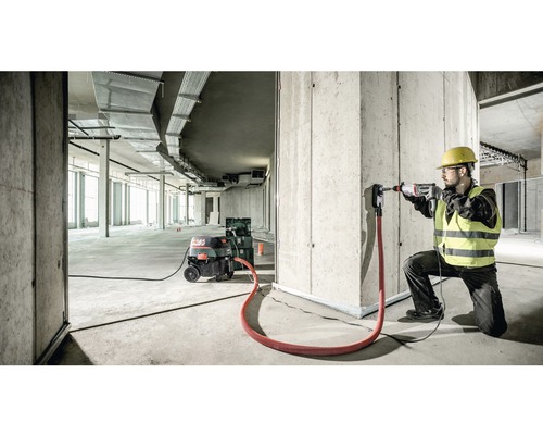 Stavebný robotník vŕta s vŕtacím kladivom otvor do betónovej steny, je pripojené odsávanie prachu.