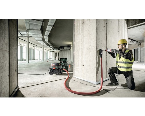 Stavebný robotník vŕta vŕtačkou otvor do betónovej steny. K dispozícii je aj priemyselný vysávač a odsávací adaptér.