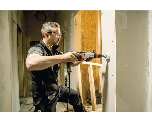 Muž vŕta vŕtačkou dieru do steny na stavenisku.