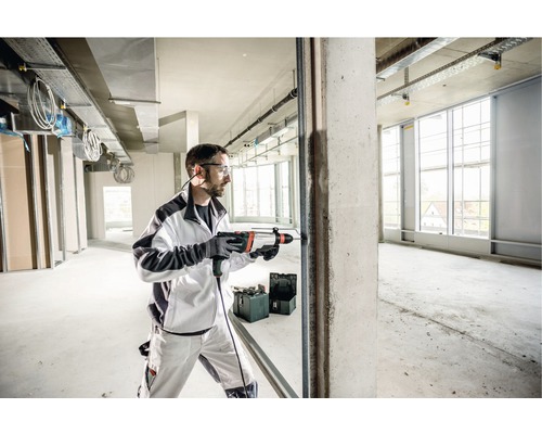 Muž vŕta vŕtacím kladivom otvor do betónovej steny v hrubej stavbe