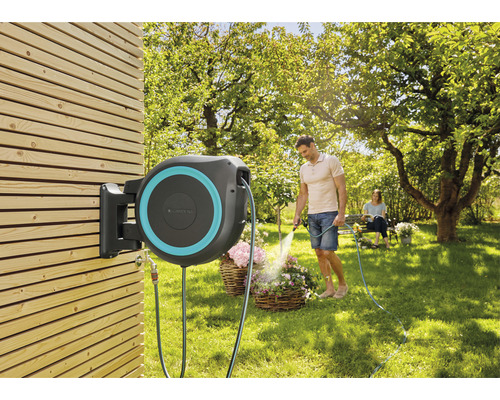 Záhradná scéna s boxom na hadicu namontovaným na drevenej stene a mužom zalievajúcim kvety