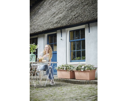 Žena sedí pri záhradnom stole pred domom s kvetináčmi a slamenou strechou.