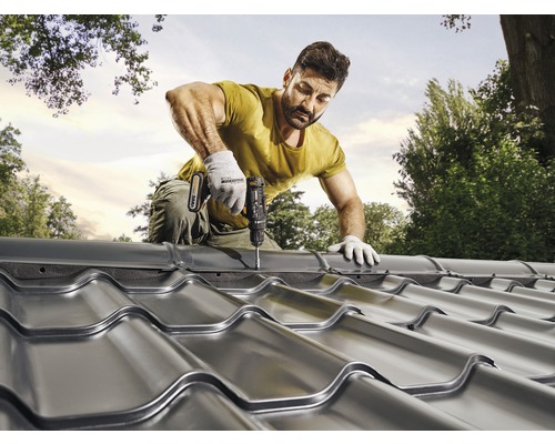 Muž pripevňuje kovové strešné tašky akumulátorovým skrutkovačom na strechu.