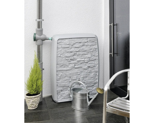 Nádrž na dažďovú vodu s kamenným vzhľadom steny, spojená so zvodom, vedľa nej krhlička a rastlina.