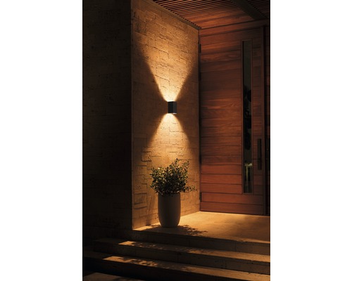 Exteriér domu s modernou nástennou lampou svietiacou hore a dole, vedľa drevených dverí a črepníkovej rastliny.