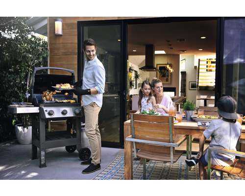 Rodina griluje vonku s plynovým grilom a dreveným jedálenským stolom.