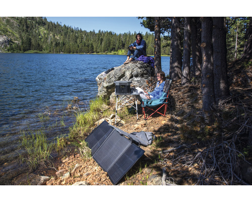 Scéna pri jazere s prenosným solárnym panelom, napájacou stanicou, stolom a kempingovými stoličkami na vonkajšie použitie.