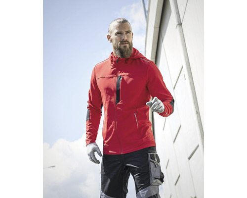 Muž s bradou, červená bunda s kapucňou, pracovné nohavice a pracovné rukavice pred fasádou budovy