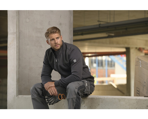 Muž v pracovnom odeve s bundou a nohavicami sedí na betóne