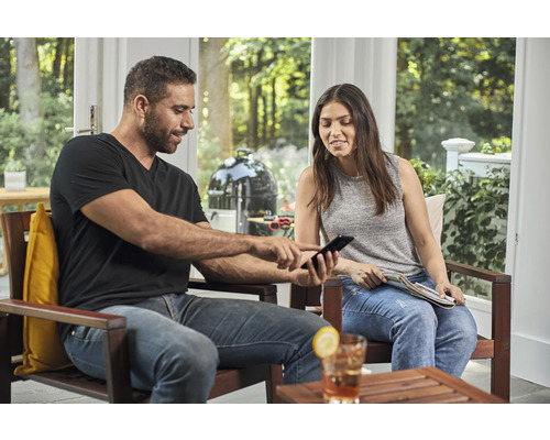 Muž a žena sedia na terase a pozerajú sa na mobilný telefón, v pozadí guľový gril.