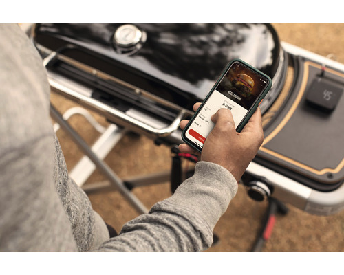 Osoba používa aplikáciu na smartfóne vedľa grilu.