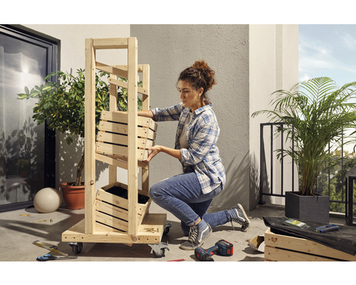 Žena montuje drevený regál s kvetináčmi na balkóne. Náradie a rastliny sú tiež vidieť.