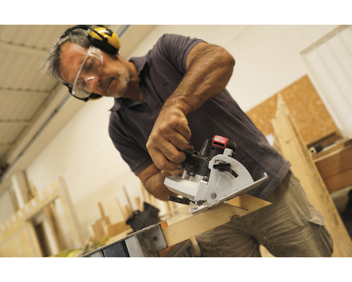 Muž pília drevenú dosku mini ručnou okružnou pílou a má pritom ochranné okuliare a chrániče sluchu.