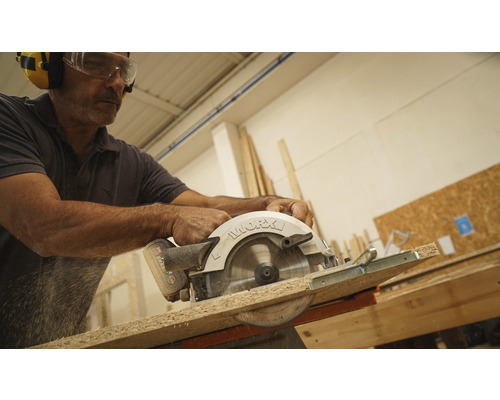 Muž pília drevenú dosku ručnou kotúčovou pílou a má na sebe ochranné okuliare a chrániče sluchu.