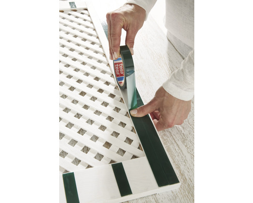 Osoba aplikuje obojstrannú lepiacu pásku na bielu drevenú mriežkovú štruktúru.