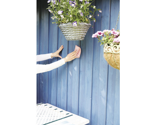 Čistenie drevenej záhradnej steny handričkou, s visiacimi košíkmi s kvetinami.