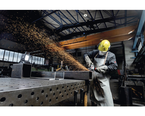 Pracovník v ochrannom odeve brúsi kov uhlovou brúskou v dielni.