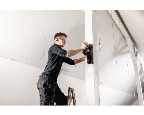 Remeselník na rebríku brúsi drevený stĺp elektrickou brúskou v podkroví.