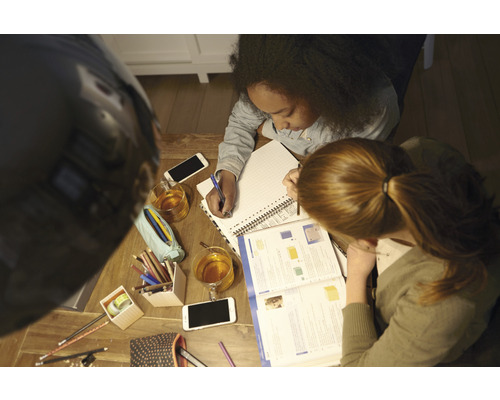 Dve osoby sa spoločne učia pri drevenom stole s poznámkovými blokmi, perami, mobilnými telefónmi a nápojmi.