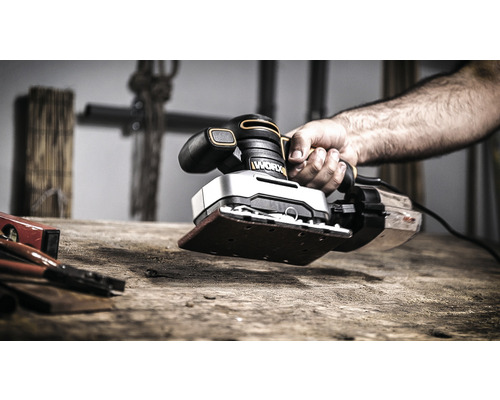 Ruka drží vibračnú brúsku značky Worx na drevenom stole.