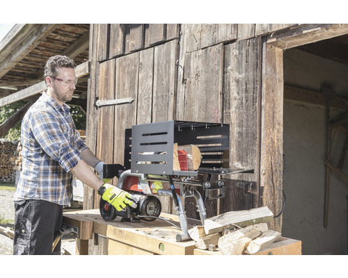 Muž obsluhuje štiepačku na drevo pred drevenou stenou. Štiepačka stojí na pracovnom stole a vedľa nej ležia kláty.