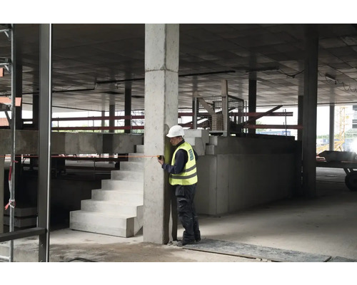 Stavebný robotník meria betónový stĺp zvinovacím metrom v hrubej stavbe.