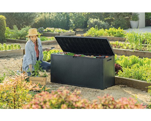 Záhradná scéna so ženou, ktorá kľačí vedľa otvoreného čierneho záhradného boxu a sadí sadenice. V pozadí je vidieť zeleninová záhrada.