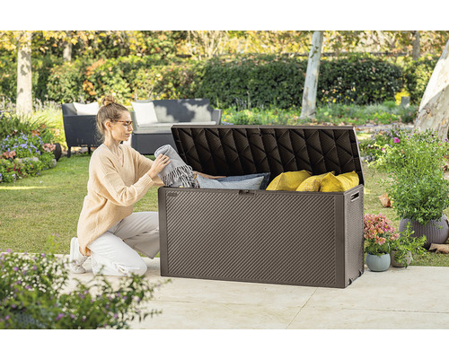 Žena ukladá deky a vankúše do hnedej záhradnej truhlice vonku.