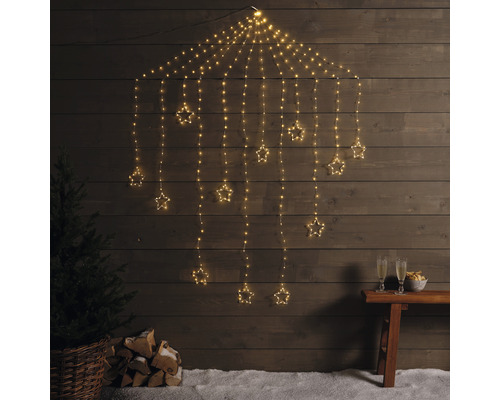 Dekoratívna svetelná reťaz s hviezdami pred drevenou stenou, so stromom, polenami a pohármi na sekt